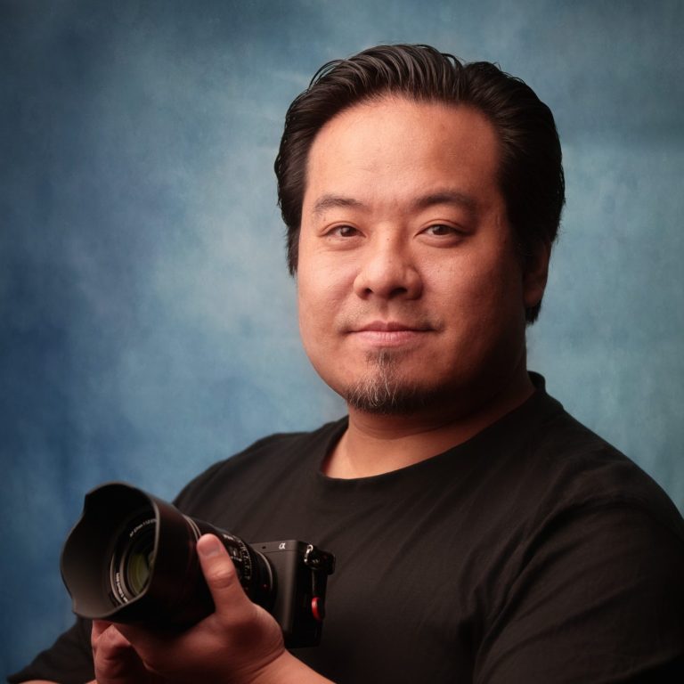 Mann mit gekreuzten Armen, lächelnd, in dunkler Kleidung vor schwarzem Hintergrund.